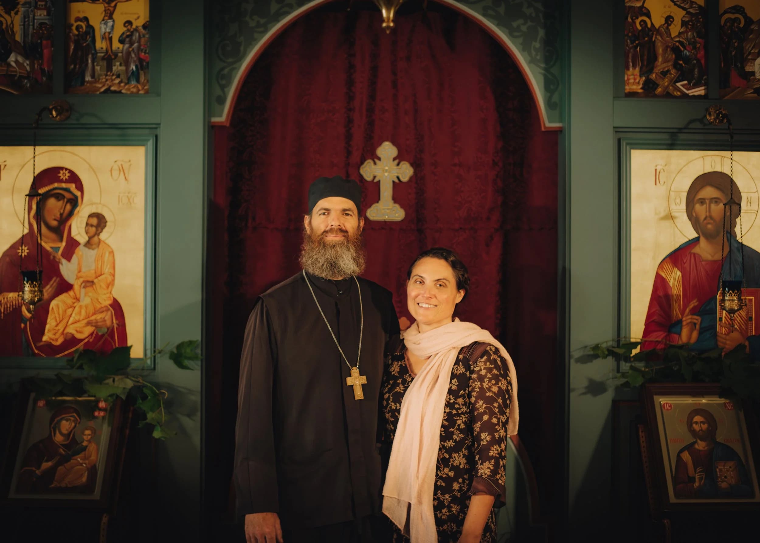 Orthodox priest and a woman standing inside the icon-filled parish church.
